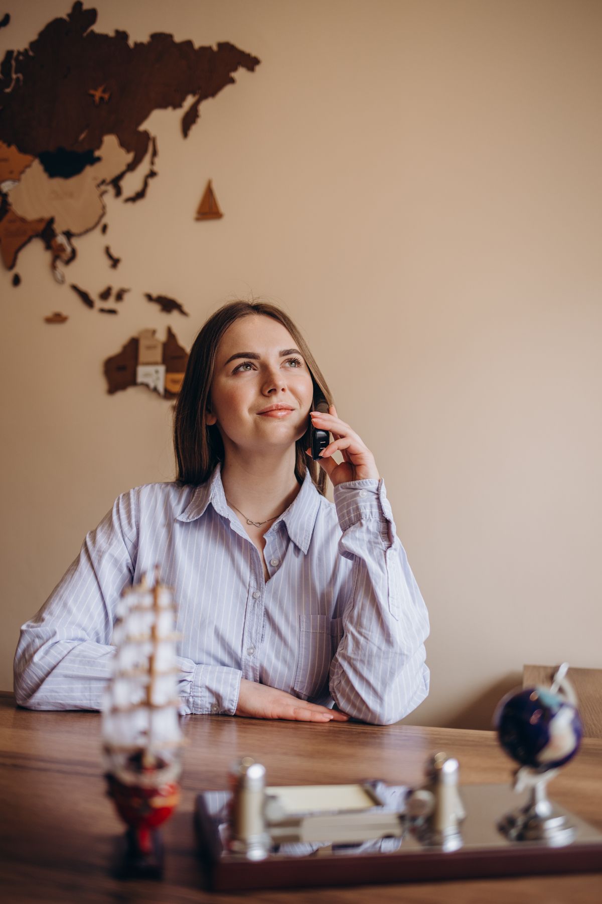 Mulher sorrindo ao telefone numa mesa de trabalho, com um mapa-múndi na parede